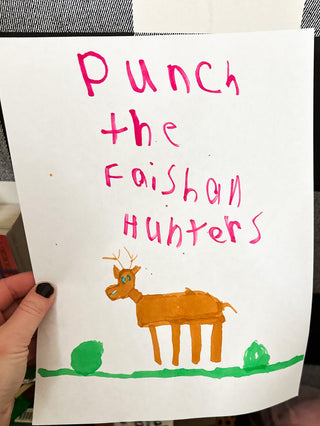 A hand holds up a white piece of paper with a child's hand-drawn illustration and text that reads 'Punch the Faishan Hunters' in pink marker, with an orange deer and green bushes below.
