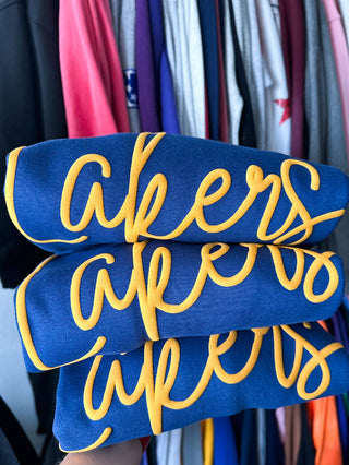 Three navy blue sweatshirts stacked together, each featuring the word 'akers' in yellow script, held in front of a background of hanging colorful clothes.